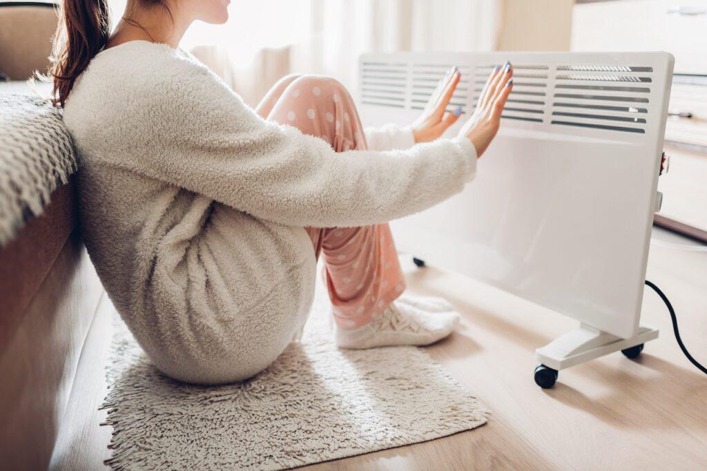 Eine Frau in warmer Kleidung sitzt auf einem Teppich und hält ihre Hände vor einen modernen Heizlüfter, um sich aufzuwärmen.