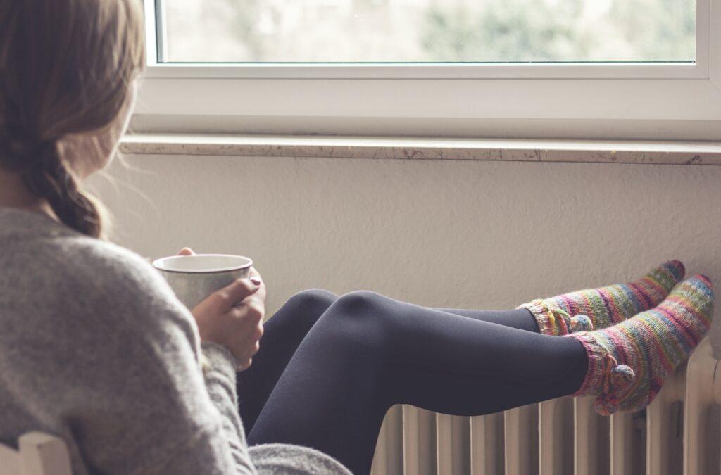 Frau sitzt mit einer Tasse Tee am Fenster, ihre Beine ruhen auf einem warmen Heizkörper, während sie Wollsocken trägt.